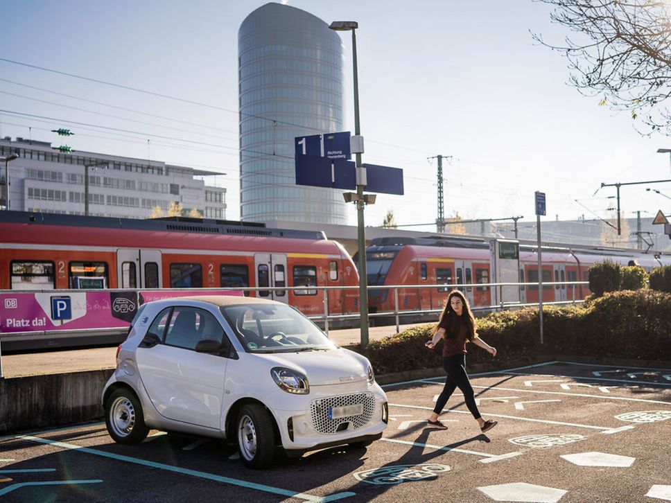 Eine Frau parkt ihren Smart auf einem Parkplatz bei einer S-Bahn Haltestelle. Im Hintergrund fährt eine rote S-Bahn ein.
