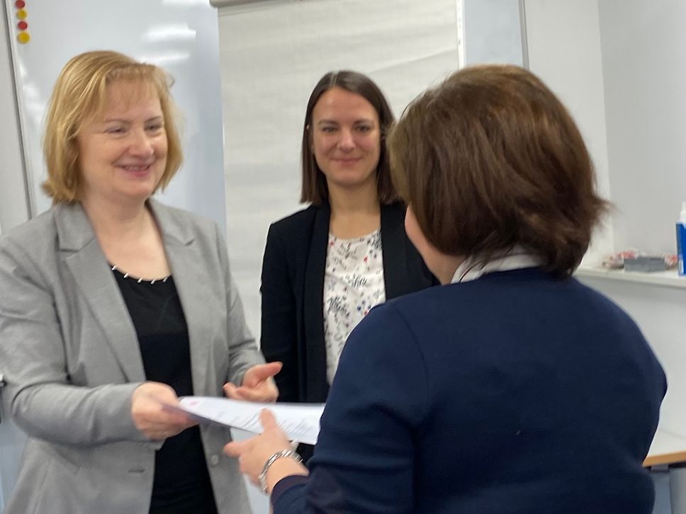 Zwei Frauen überreichen einer weiteren Frau eine Urkunde