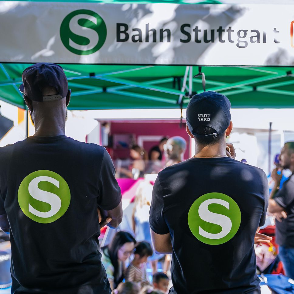 Zwei Mitarbeiter der S-Bahn Stuttgart stehen mit dem Rücken zur Kamera und das S-Bahn-Symbol ist auf dem Rücken erkennbar.